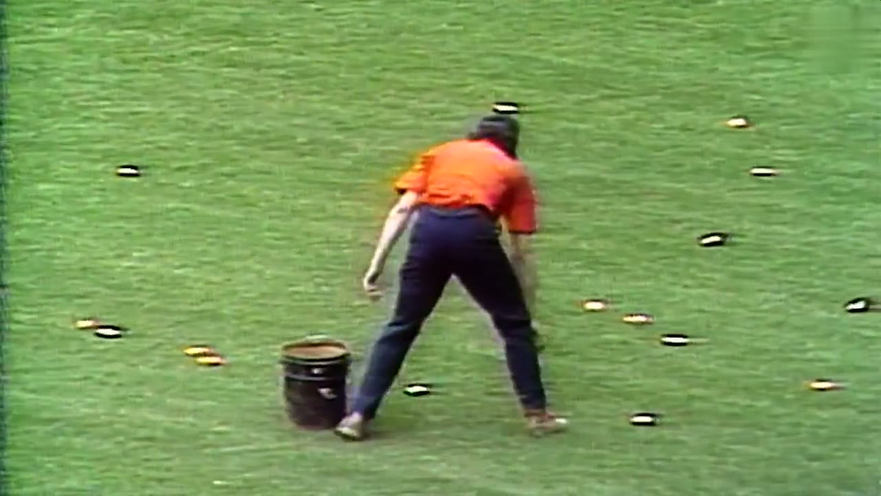 A grounds crew member with a bucket picking up dozens of orange REGGIE! Bar wrappers scattered across the Yankee Stadium outfield in 1978