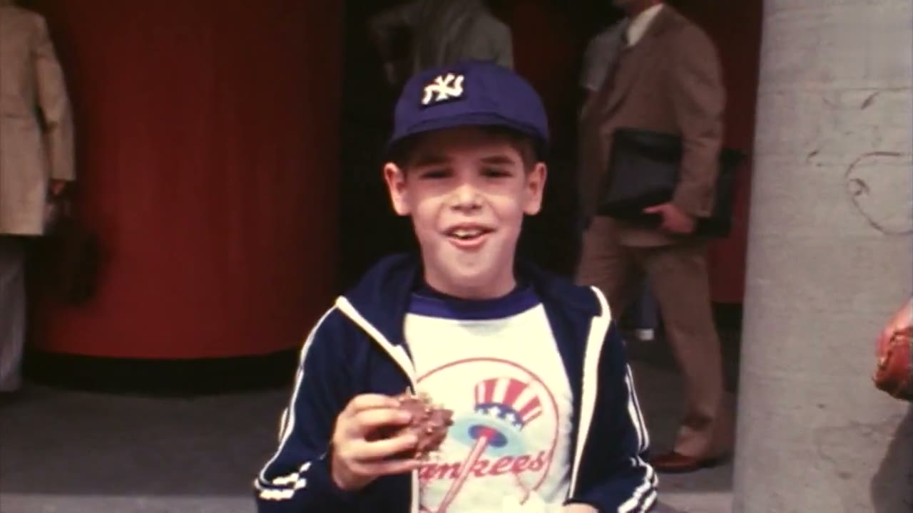 A young fan in a Yankees jacket and cap smiling as he eats a REGGIE! Bar at the ballpark in 1978