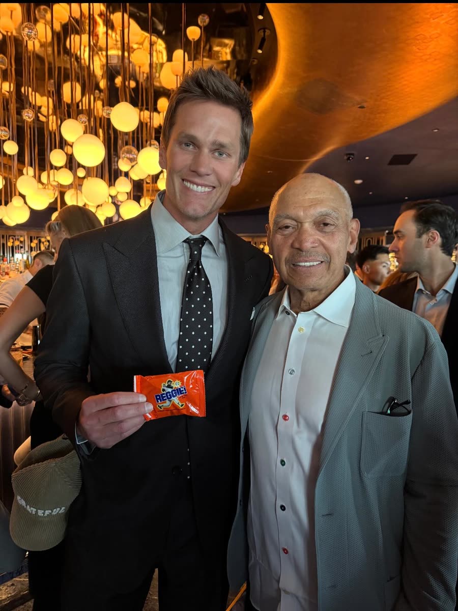 Tom Brady holding a REGGIE! Bar while standing next to Reggie Jackson at an evening event