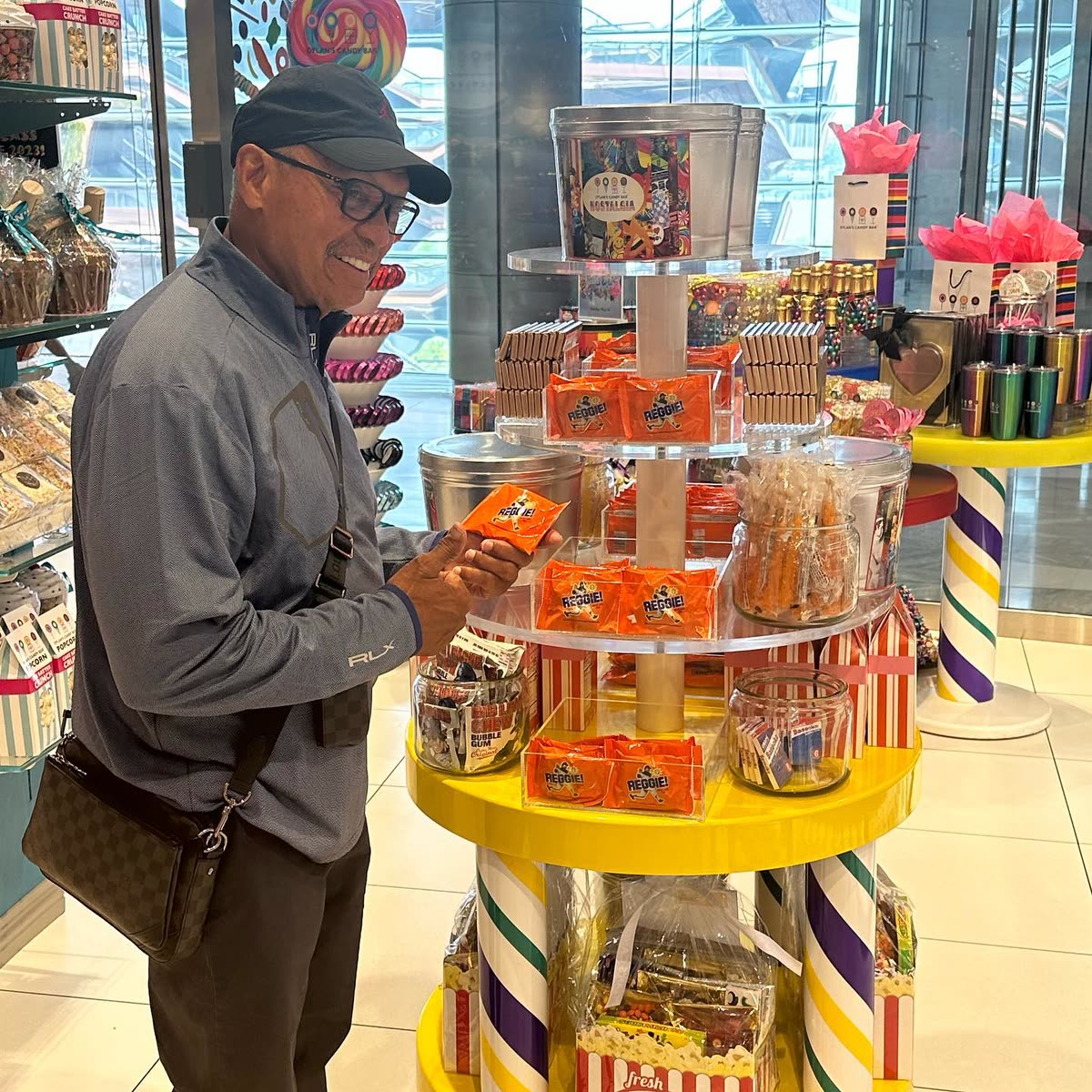 Reggie Jackson smiling as he holds a REGGIE! Bar beside a tiered candy store display stacked with them